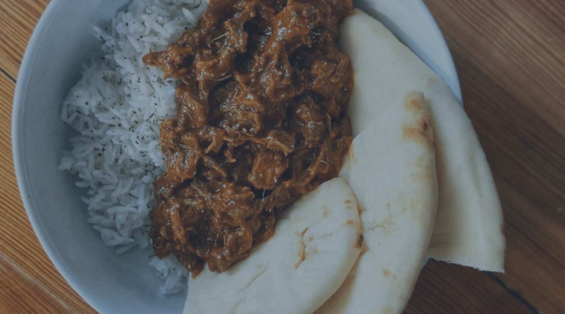 goat curry in a white bowl with rice and naan