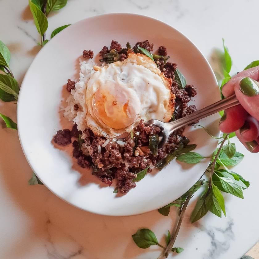 White bowl with goat pad era pao topped with a fried egg, fork is holding some of the ground meat above the bowl