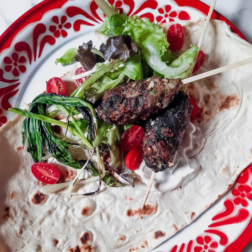 Kofta kabobs with lettuce, tomato, yogurt sauce on a pita on a red and white plate