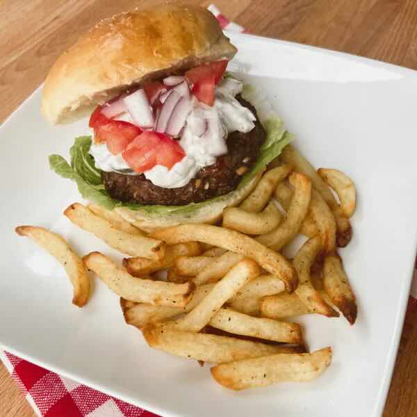 Greek goat burger with tomatoes and onions and fries on a white plate. 